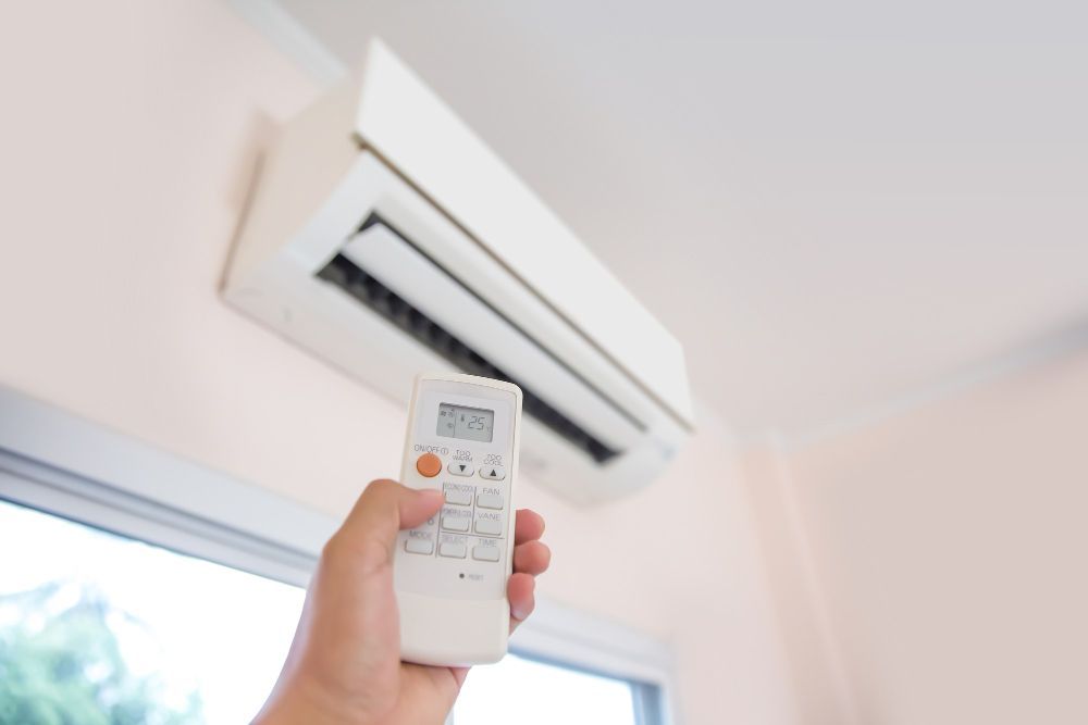 Hand holding a remote pointed at a wall-mounted air conditioner in a bright room