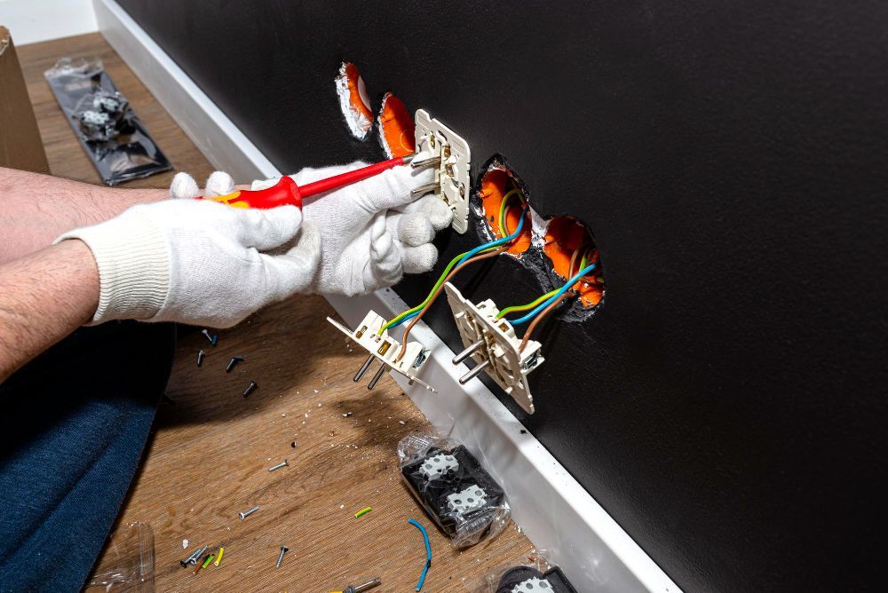 Gloved hands wiring an electrical outlet in a wall, with exposed cables and tools on the floor.