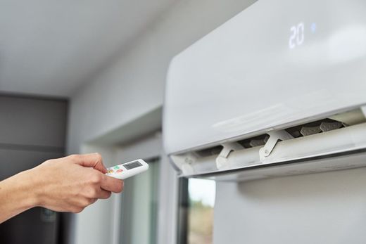 Hand holding a remote pointed at a wall-mounted air conditioner, with the display showing 20°C.