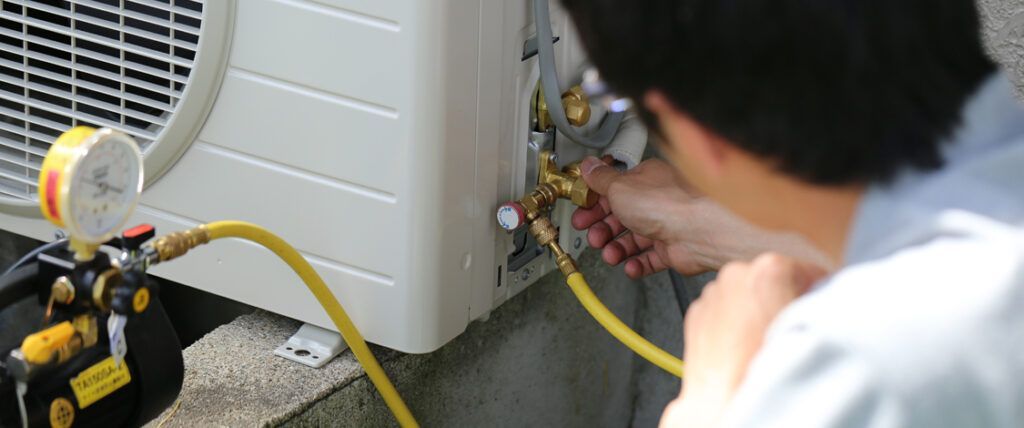 Technician connects a yellow hose to an outdoor HVAC unit while another person watches.
