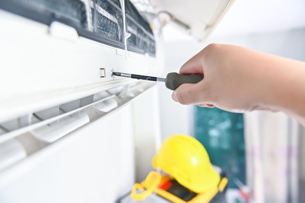 Hand tightening an air conditioner vent filter with a screwdriver, yellow hard hat below