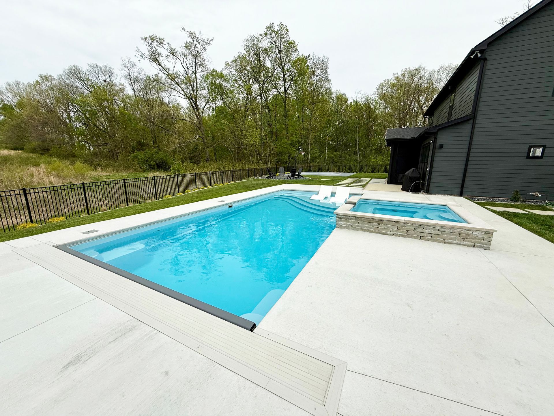 A rectangular swimming pool with an attached stone hot tub on a concrete patio next to a dark-sided house and trees.