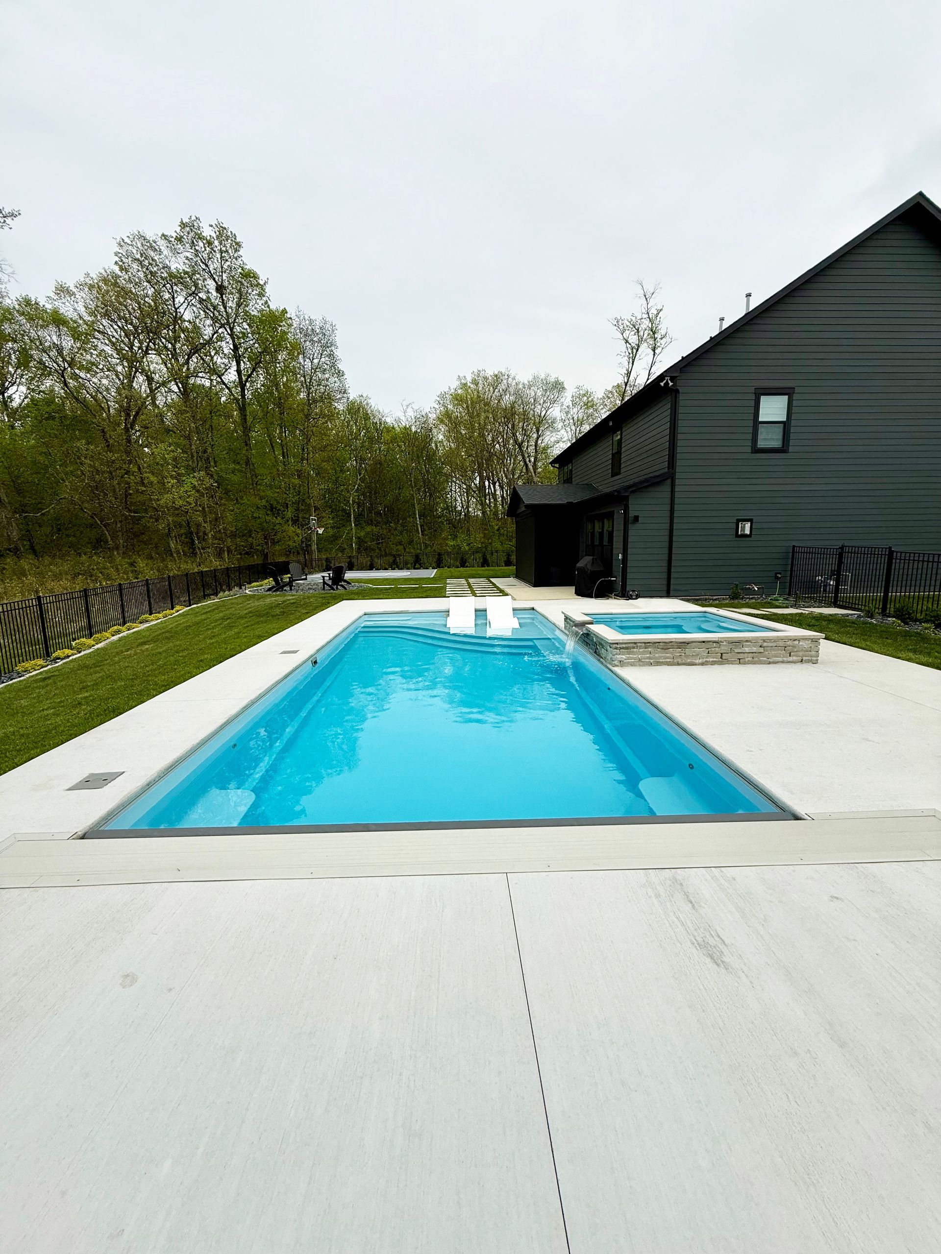 A rectangular blue swimming pool and attached hot tub sit on a concrete patio beside a modern dark-sided home.