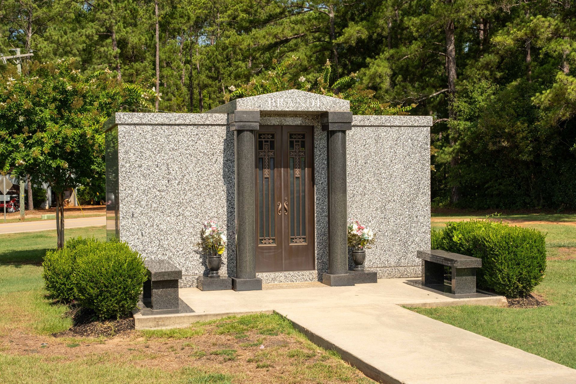 Grave-site mausoleum with a granite facade, a dark wooden door, and two pillars. A bench and bushes sit in front.
