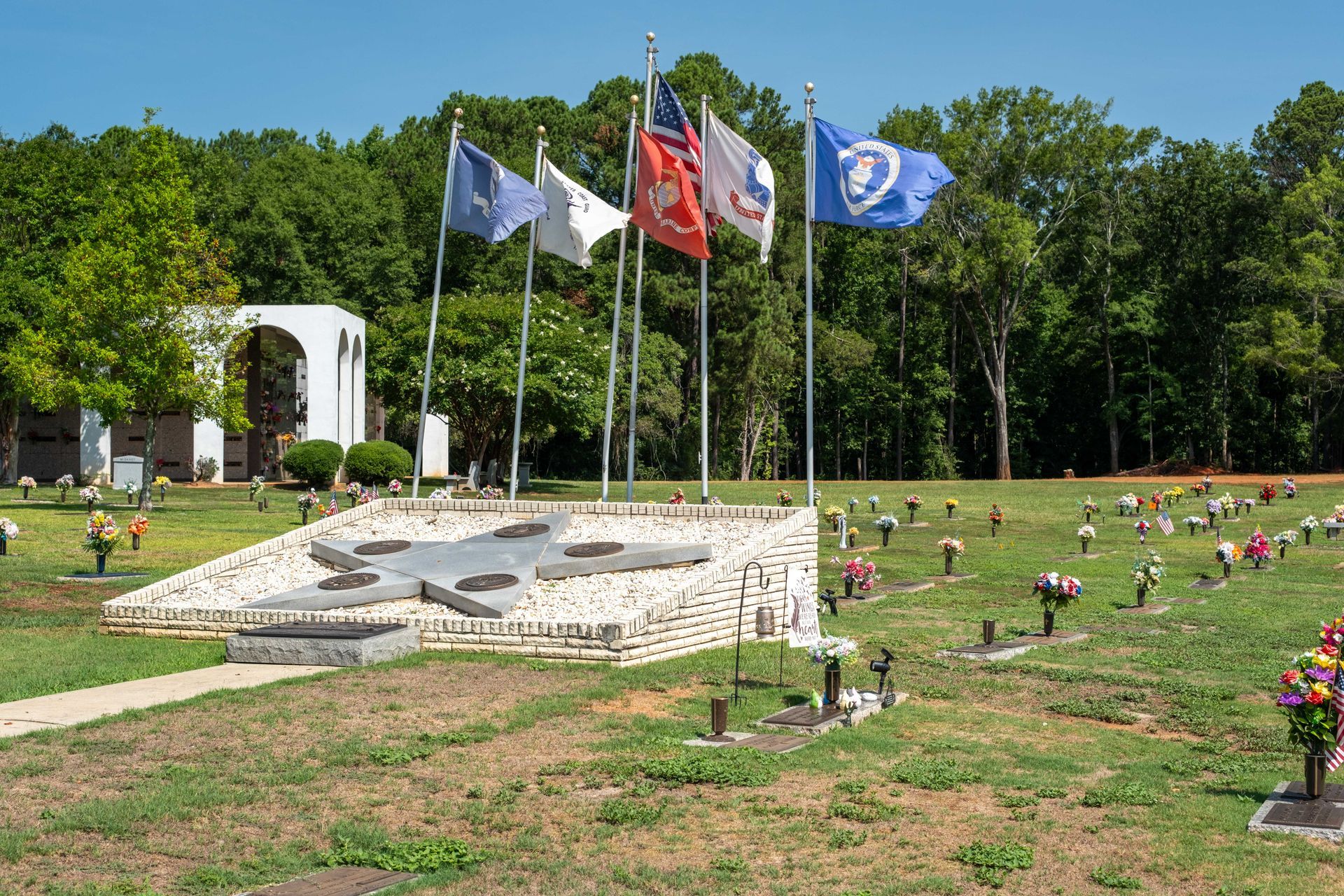 Memorial with flags waving, graves, and white structure against a green, wooded backdrop.