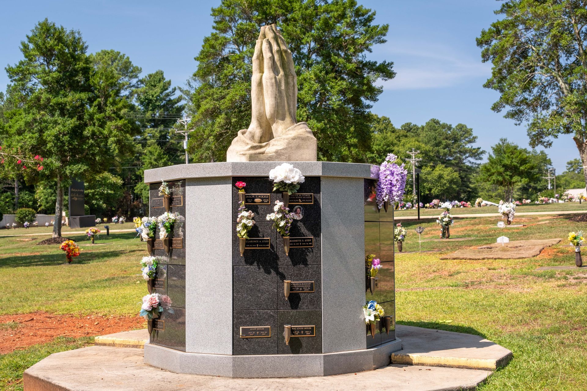 A mausoleum in a cemetery with a statue of praying hands. Flowers adorn the crypts.