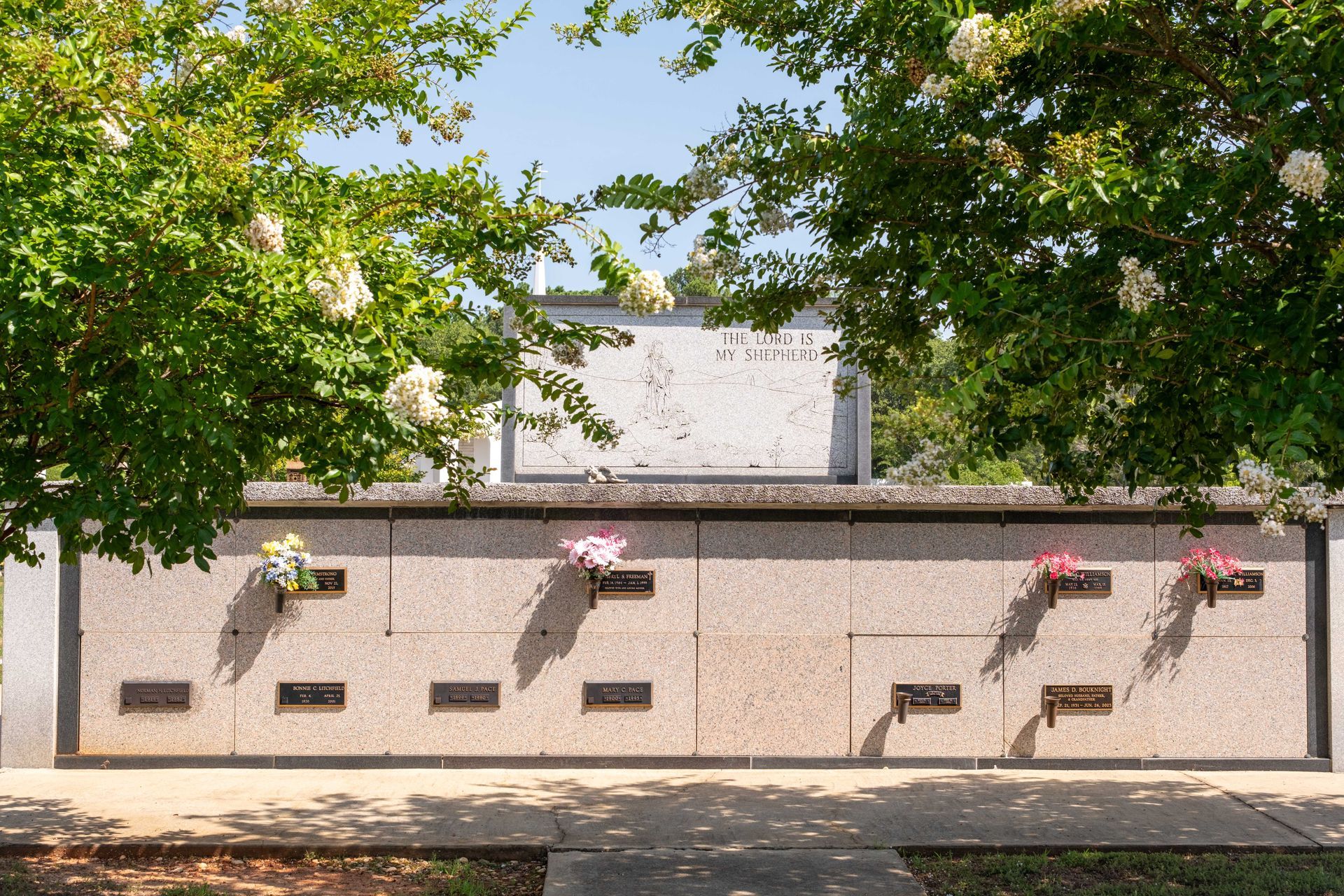 A columbarium with multiple niches and flower vases. Above is a large stone monument, with trees on both sides.