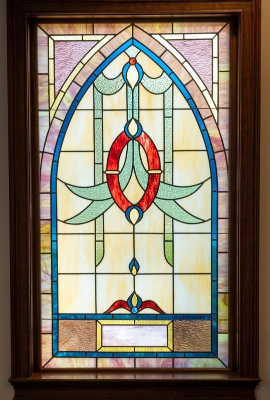 Stained glass window with an arched top. Features teal, red, yellow, and clear glass set in a dark brown wooden frame.