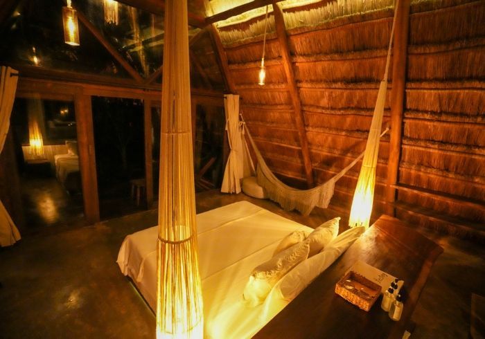 Habitación en una cabaña con techo de paja, iluminación cálida, cama con dosel y cortinas colgantes.