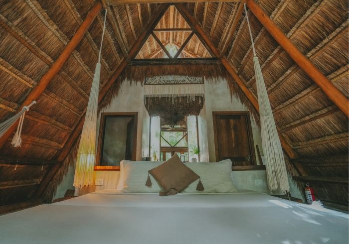 Habitación con techo de paja, cama, sábanas blancas, vigas de madera y cabecero con ventana.