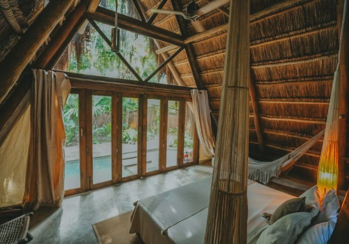 Interior de cabaña con techo de paja, camas tipo hamaca, vigas de madera y grandes ventanales con vistas a la vegetación.