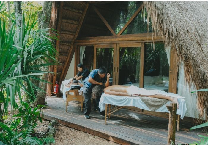 Dos personas dan un masaje en una terraza de madera junto a una cabaña tropical rodeada de exuberante vegetación.