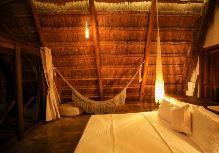 Interior acogedor de una cabaña con techo de paja, hamaca, cama de madera y cálida iluminación ámbar.