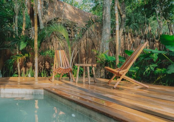 Terraza de madera con dos tumbonas junto a una piscina de color turquesa en un exuberante jardín tropical.