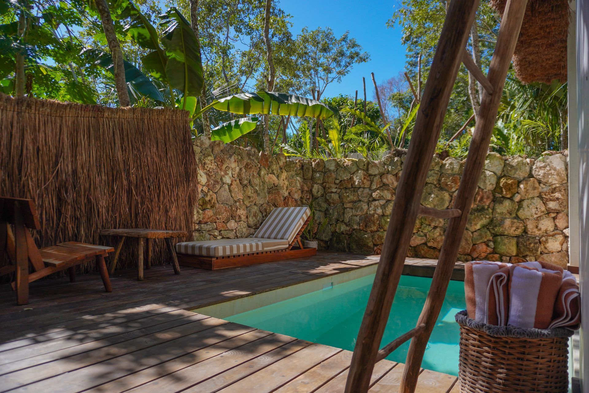 Piscina tropical a la sombra con tumbonas, muro de piedra y cesta de mimbre sobre una terraza de madera.