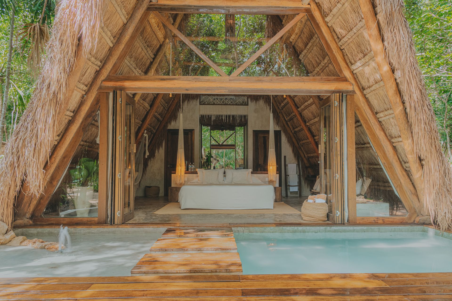 Villa al aire libre con techo de paja, terraza de madera, piscina y salón interior con vistas a una exuberante vegetación.