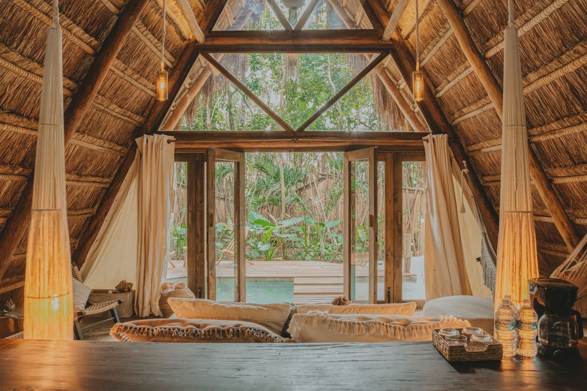 Interior de una cabaña de bambú, una gran ventana, techo tejido y cojines en el suelo con vistas a la vegetación verde.
