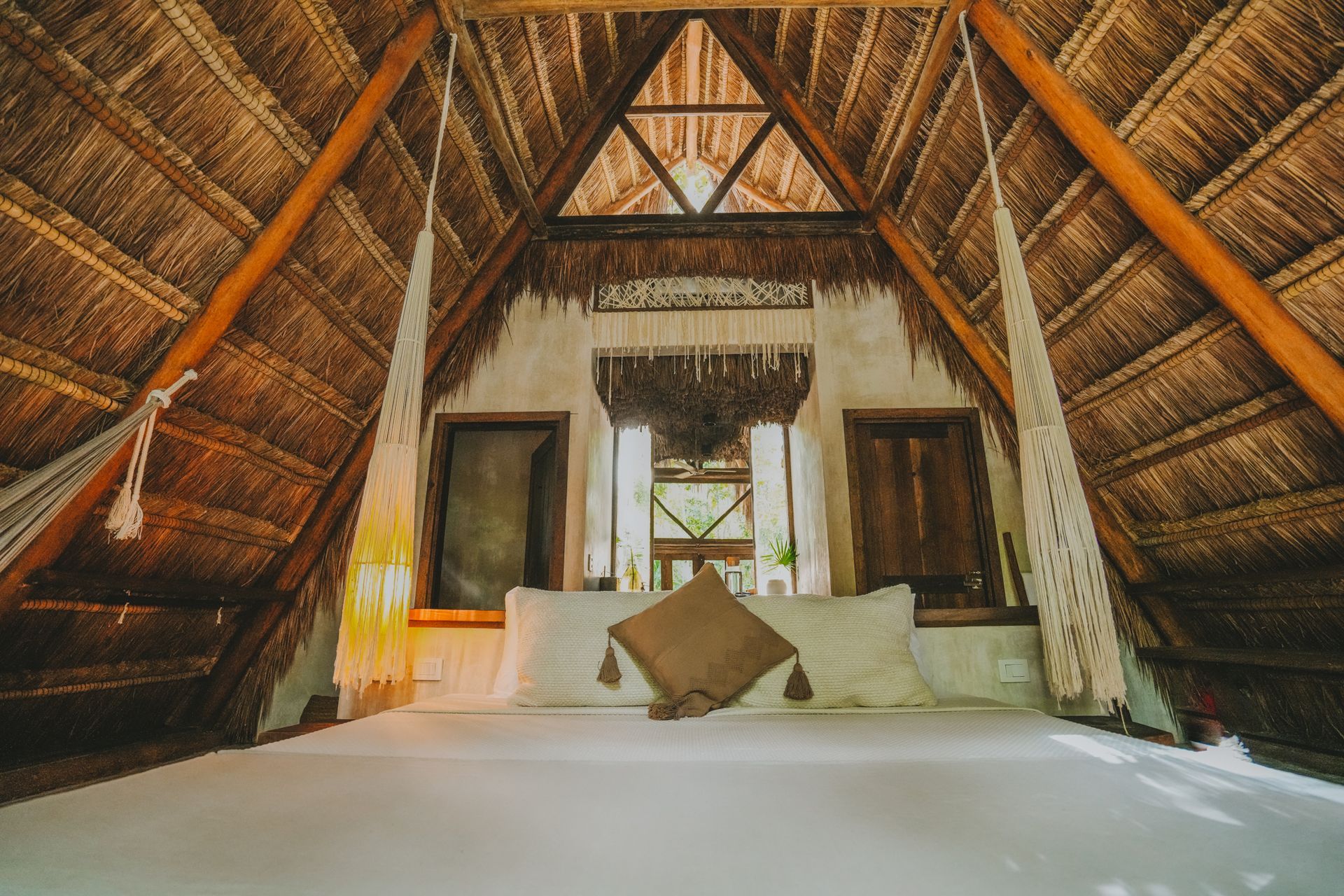 Habitación con techo de paja, cama y almohadas blancas, cálidas vigas de madera y ventanas abiertas al fondo.