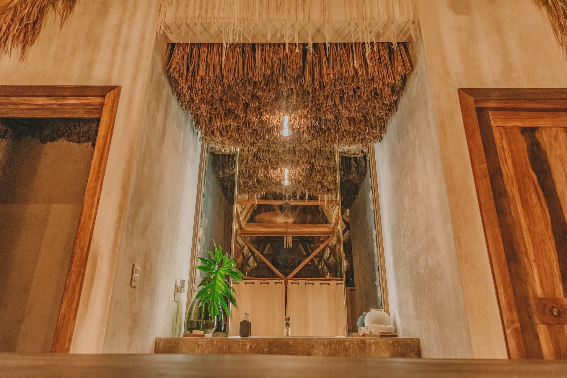Elegante recibidor con techo de paja, lámparas colgantes, puertas de madera y una pequeña planta en una repisa.