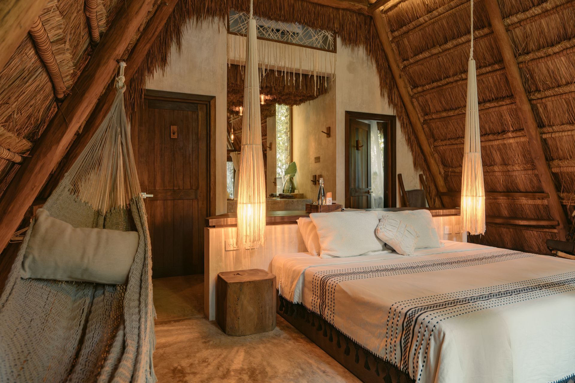 Acogedora habitación con techo de paja, cama de madera, lámparas colgantes e iluminación ambiental cálida.