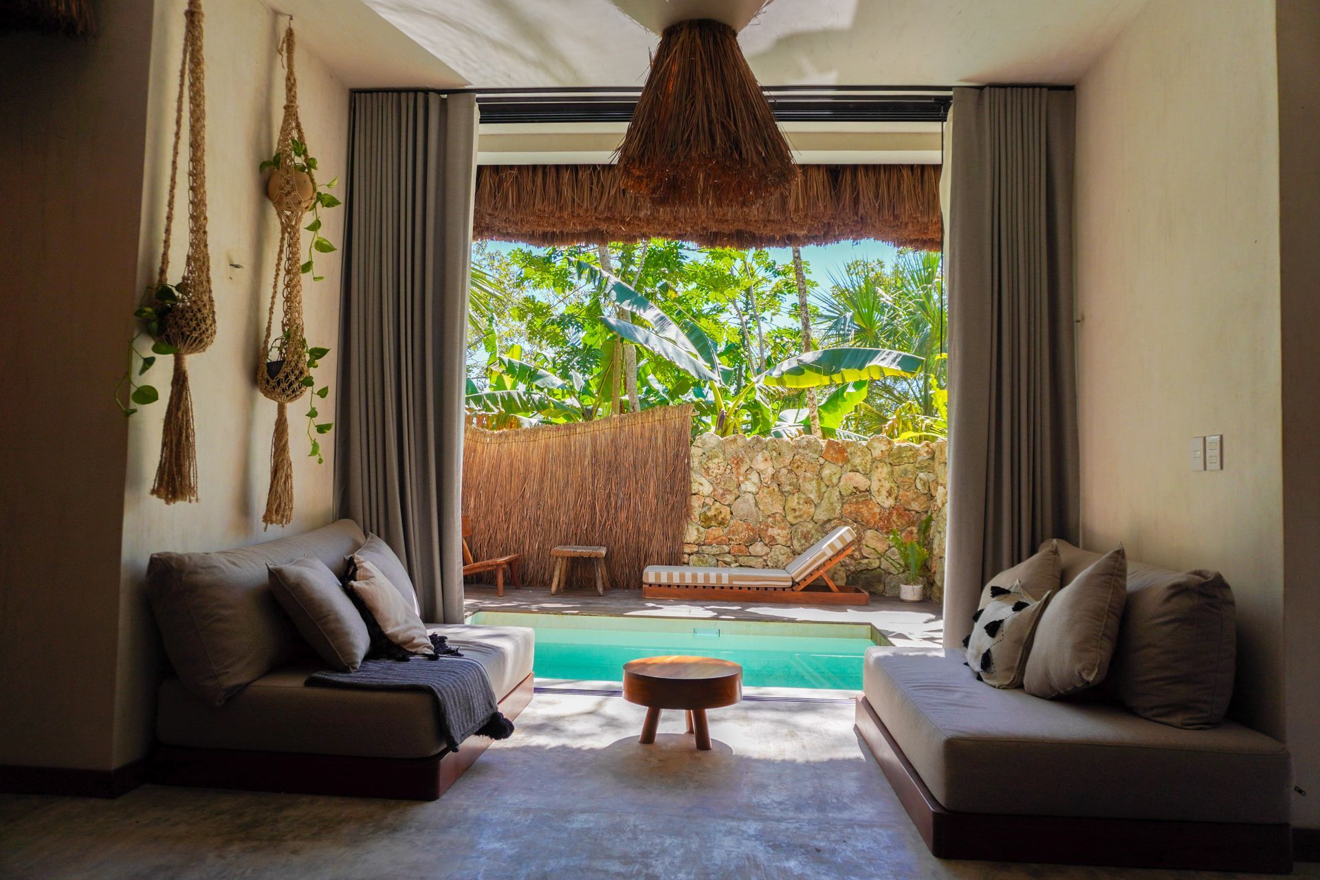salón con dos sofás grises frente a una pequeña mesa, con acceso a un patio junto a la piscina y exuberante vegetación.