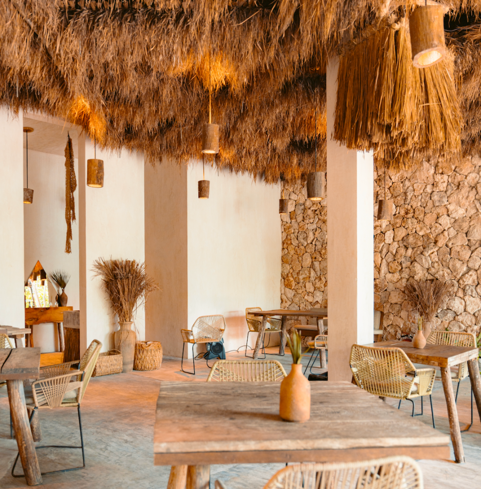 Cafetería rústica al aire libre con sillas de mimbre, mesas de madera y techo de paja.