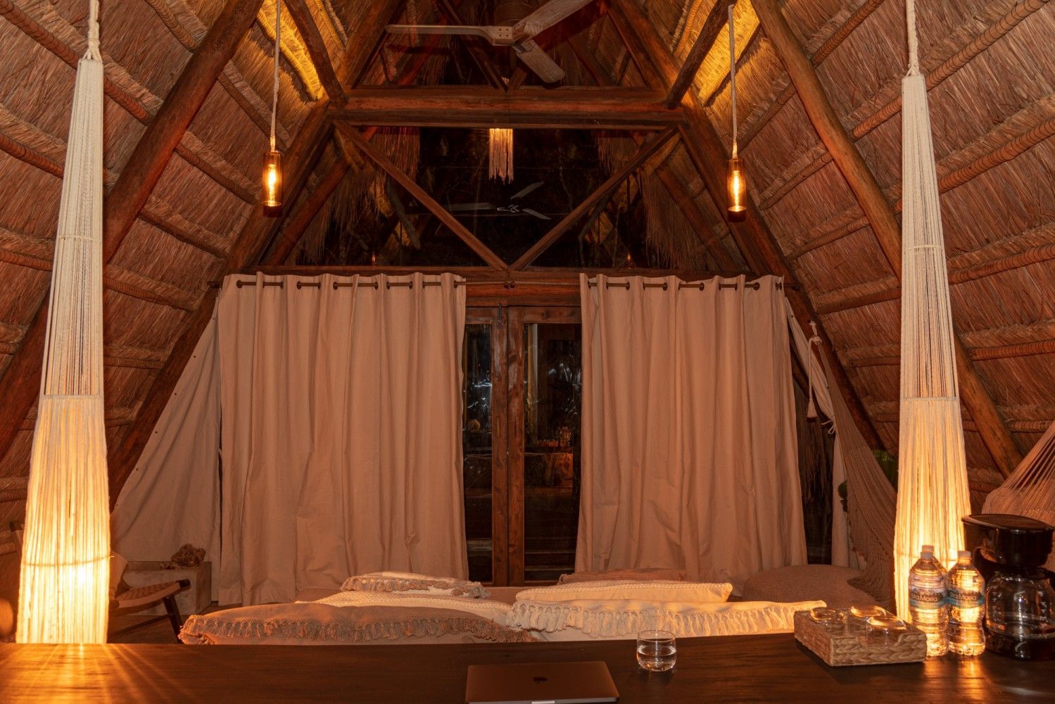 Acogedora habitación con techo de paja, cama, cortinas drapeadas y cálida iluminación ambiental.