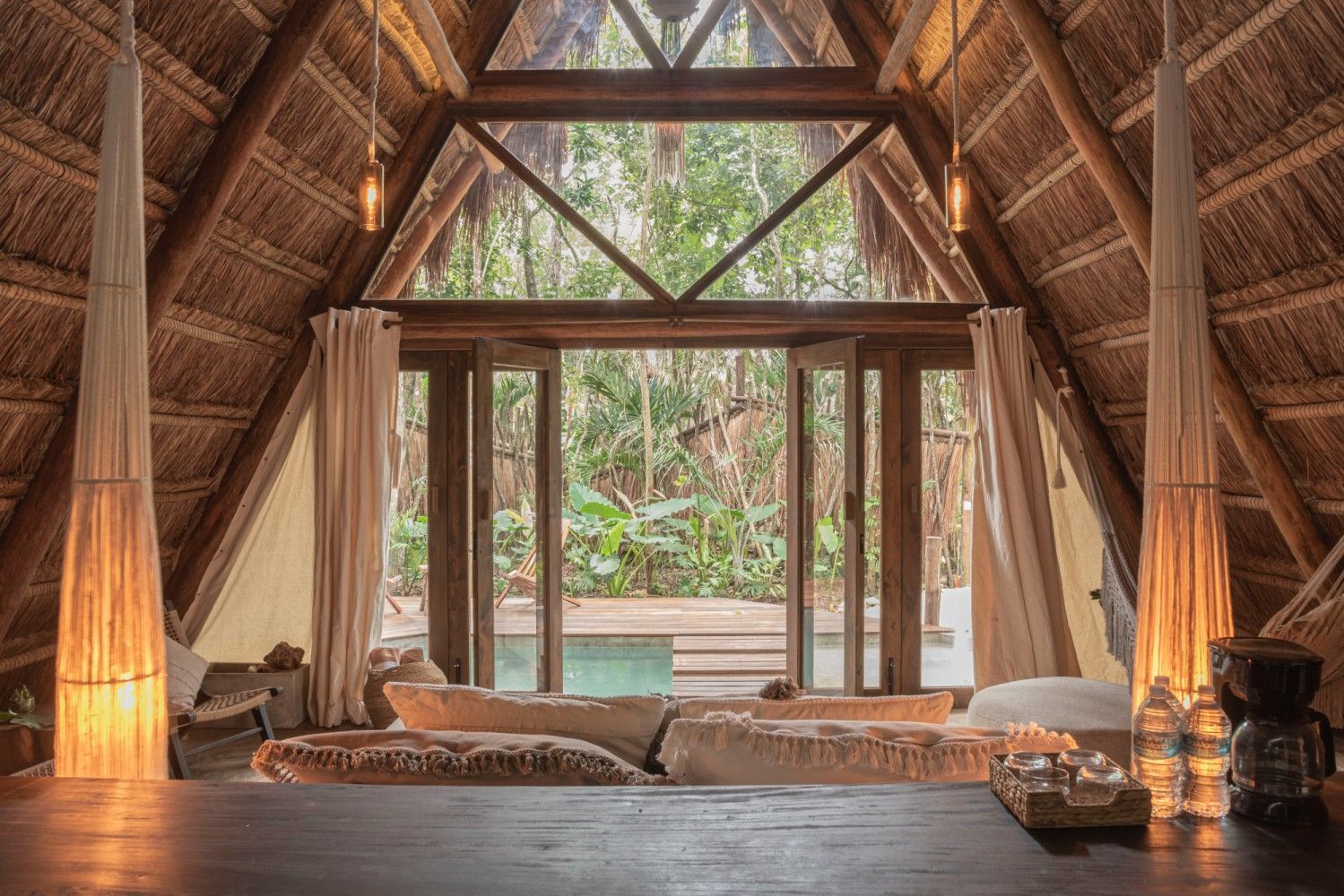Acogedora cabaña con techo de paja, cama, cortinas, lámparas cálidas y vistas a una exuberante vegetación exterior.