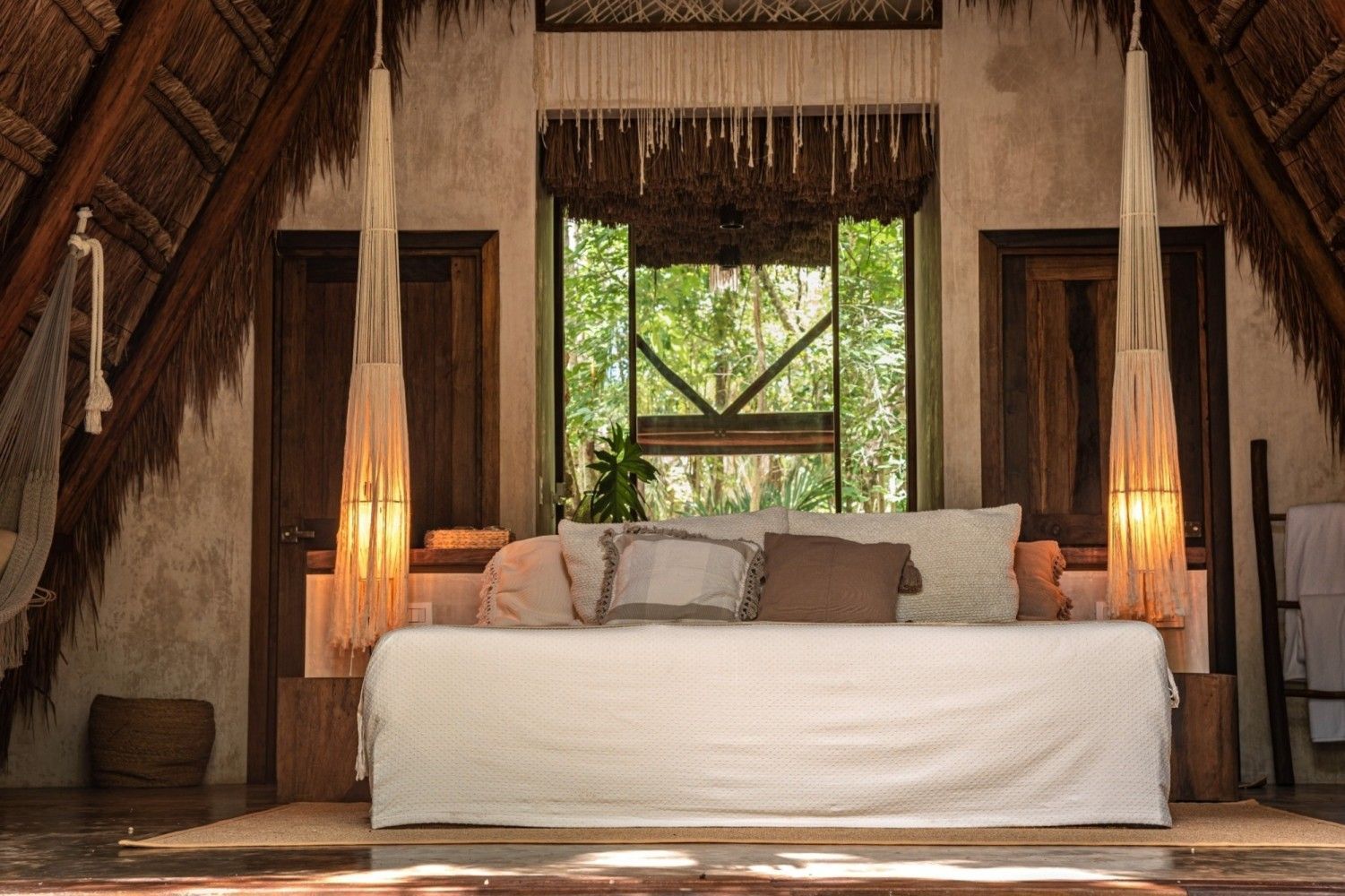 Habitación rústica tropical con una cama blanca, vigas de madera y una ventana con vegetación en el exterior.