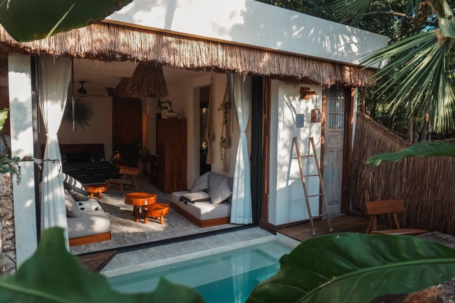 Villa tropical al aire libre con piscina, sofás blancos y muebles de madera cálida bajo un techo de paja.
