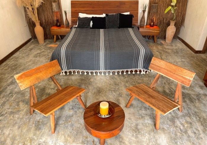 Habitación rústica con una cama de madera, una manta azul y dos sillas de madera alrededor de una mesita con una vela.