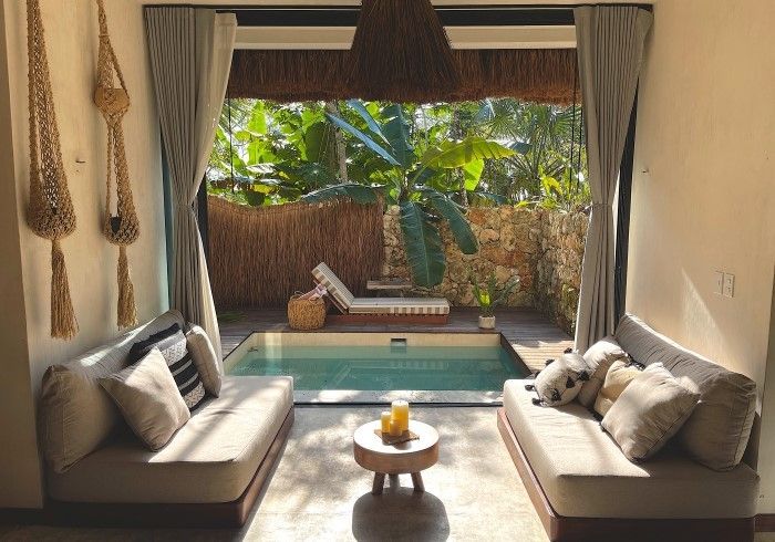 Luminoso salón con cortinas abiertas, dos sofás y vistas a una piscina y un jardín tropical.