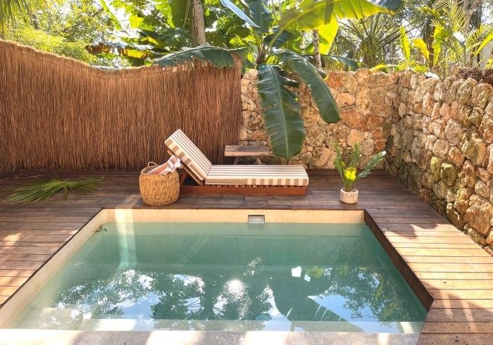 piscina en el patio trasero con terraza de madera, tumbona, cesta de mimbre y muro de piedra bajo la cálida luz del sol.