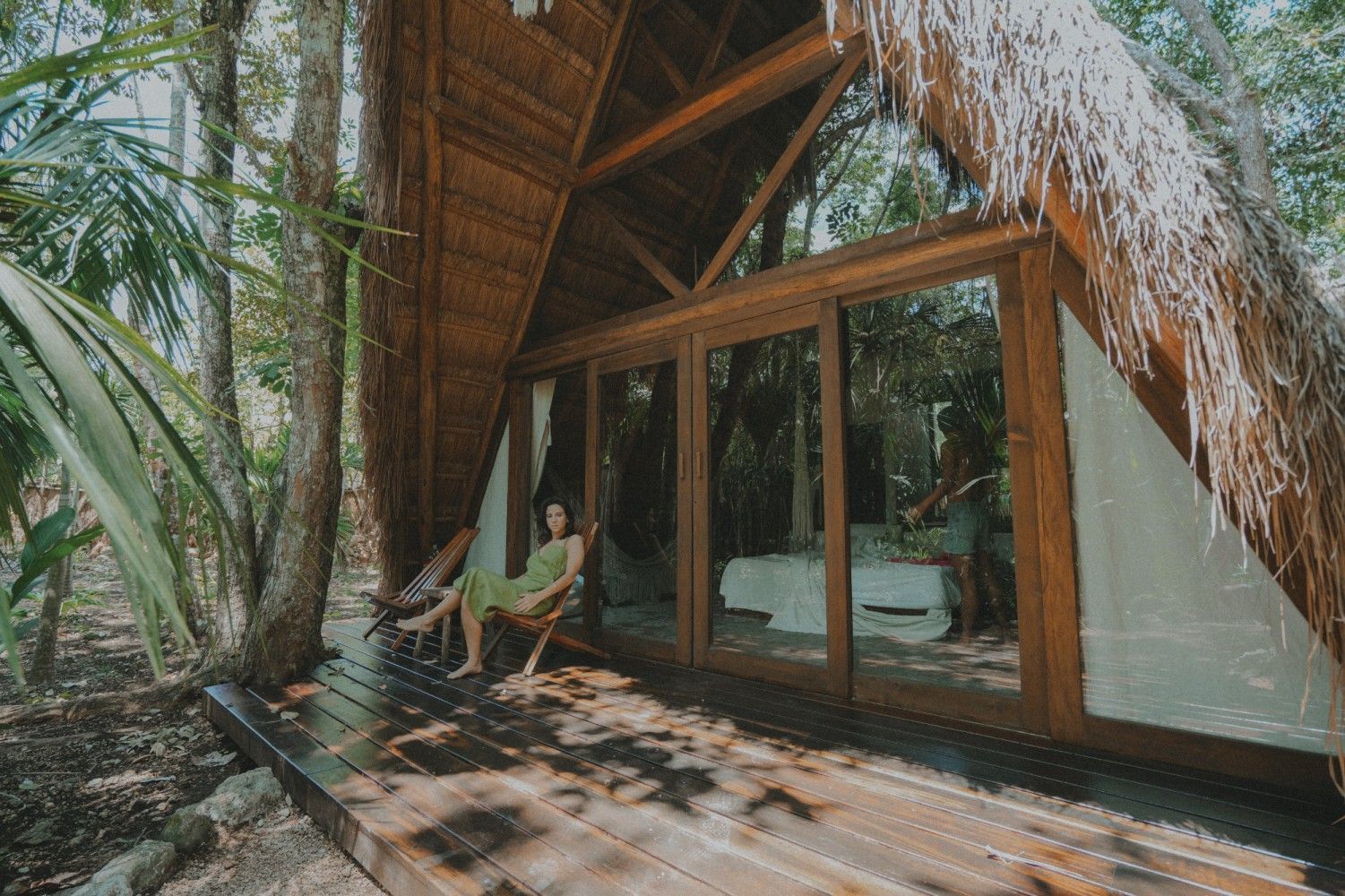 Terraza de cabaña tropical con techo de paja, sillas de madera, puertas de cristal y exuberante vegetación en las cercanías.