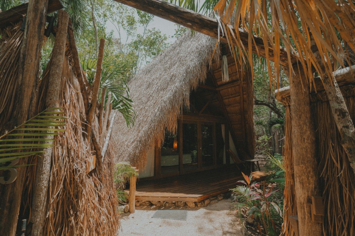 Entrada a una cabaña rústica con techo de paja, enmarcada por hojas de palmera y vegetación tropical.