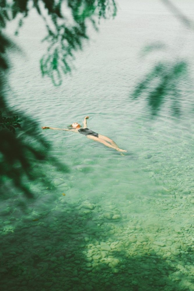 Persona flotando en agua verde clara, vista desde arriba, con sombras oscuras de hojas a su alrededor.