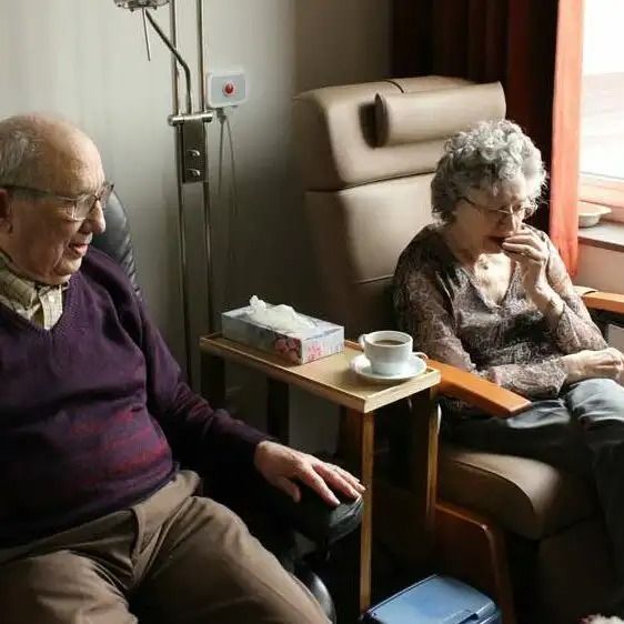 Two people sitting in armchairs beside a small table with a cup and tissue box, near a window.