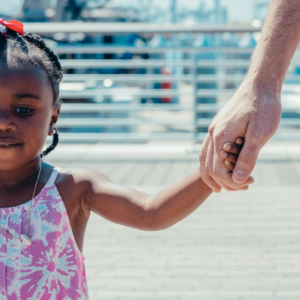 A child holding an adult’s hand on a sunlit boardwalk, with a blue railing in the background