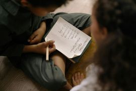 Person writing in a notebook while seated on a floor, with another person nearby.