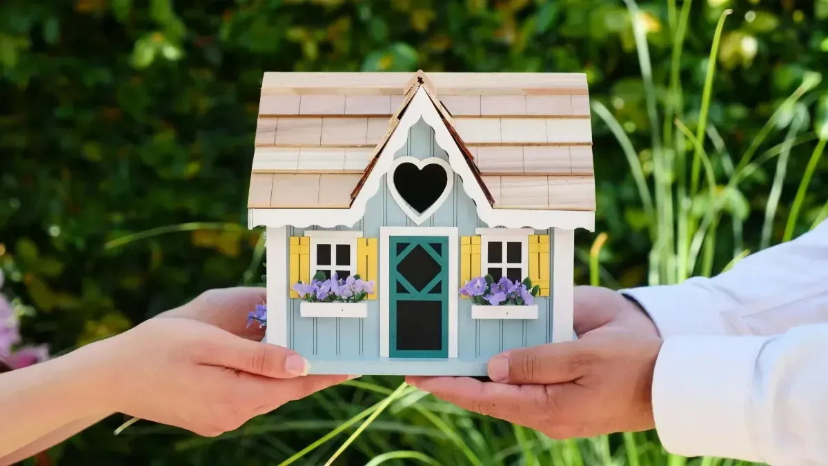 Hands exchanging a small white miniature house with a heart-shaped window outdoors