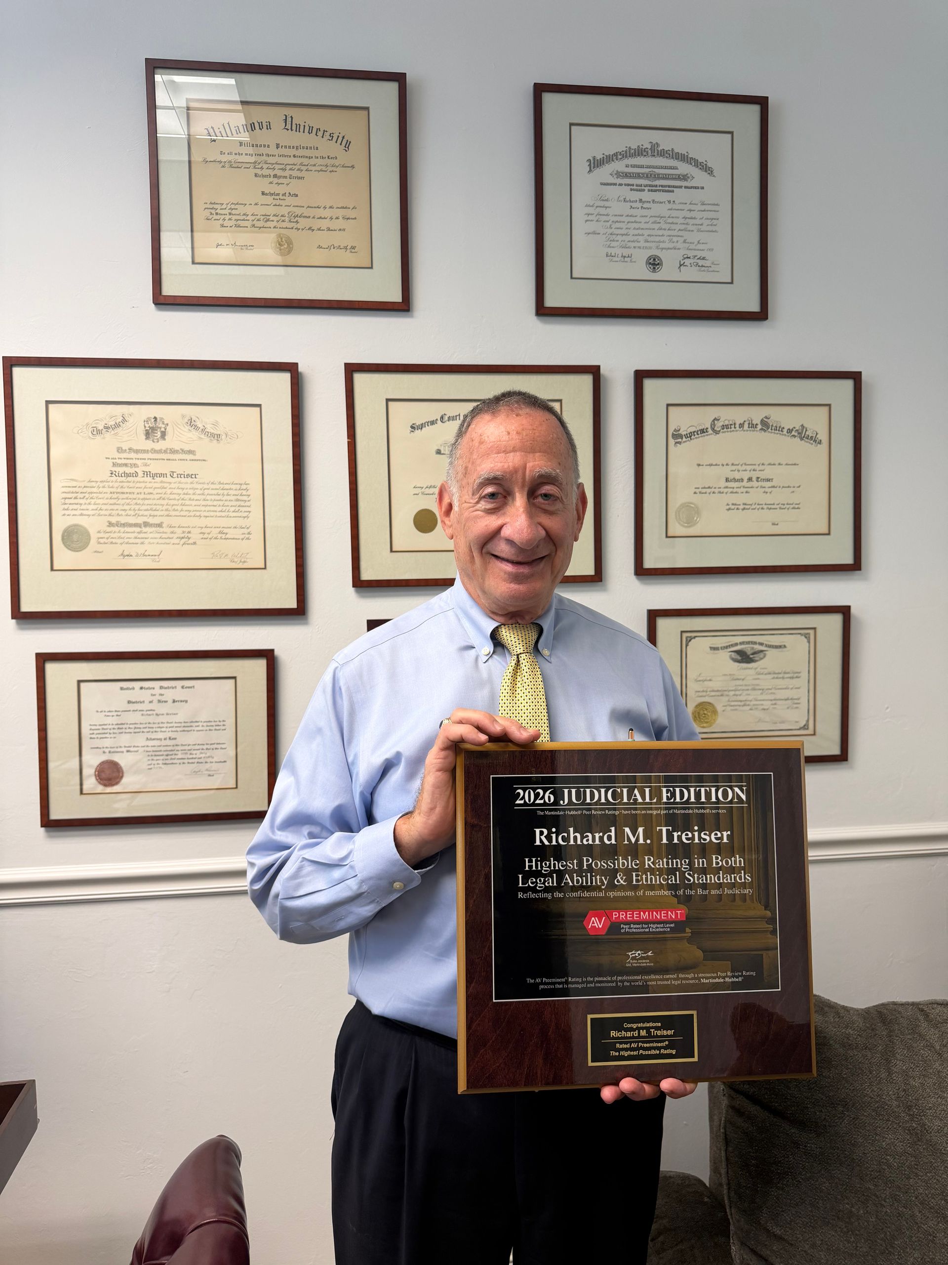 Man smiling in an office, holding a large award plaque with framed certificates on the wall behind him