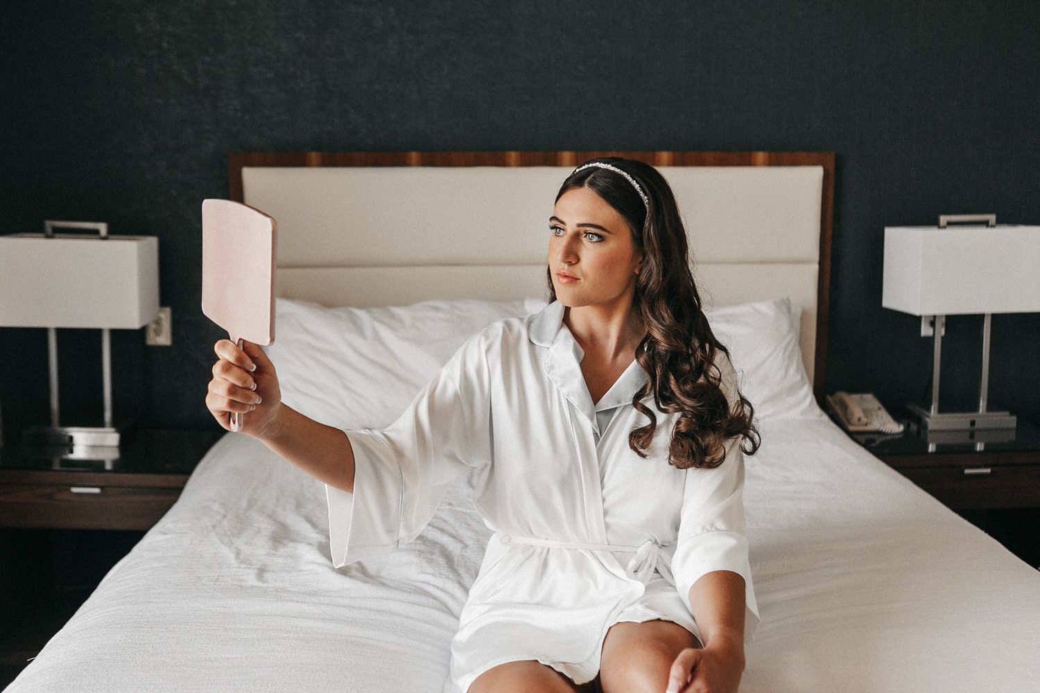 A woman is sitting on a bed looking at herself in a mirror. Orange county hair and makeup artist.