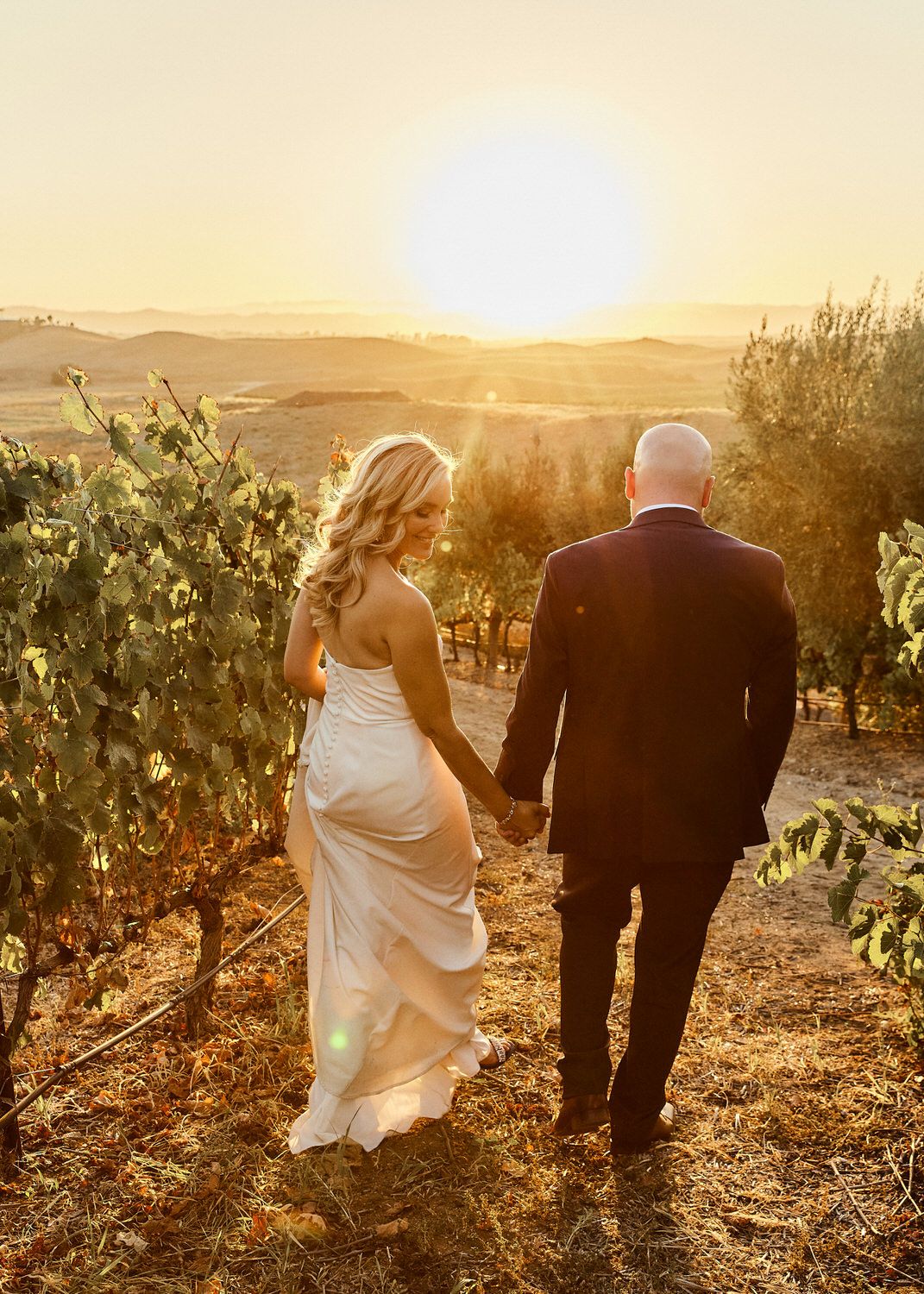 A bride and groom are walking through a vineyard at sunset holding hands. Orange county hair and makeup artist.
