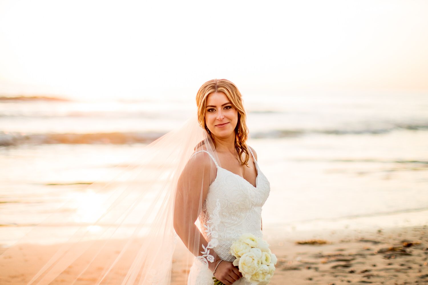A bride in a wedding dress is standing on the beach holding a bouquet of flowers.