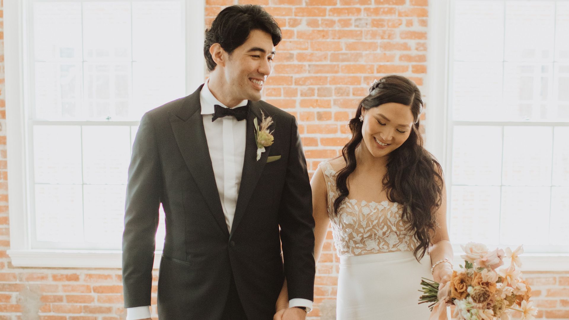 A bride and groom are holding hands and smiling in front of a brick wall.