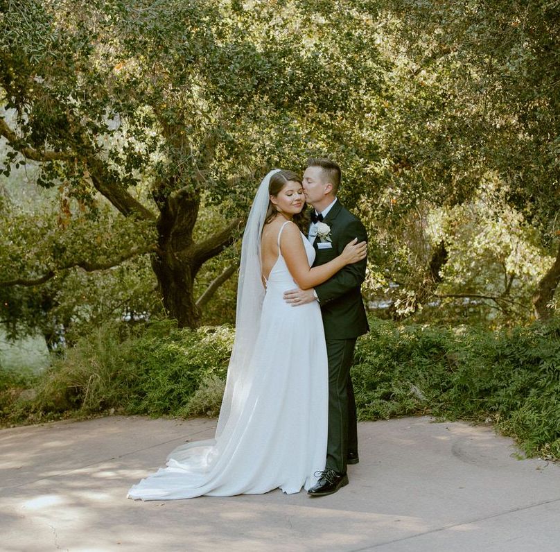 A bride and groom are kissing under a tree on their wedding day. Orange county hair and makeup artist.