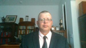 Person in a dark suit and tie seated indoors in front of bookshelves and framed pictures.