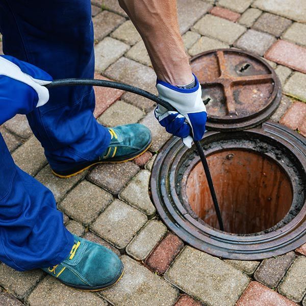 Worker in blue gloves and shoes using a hose to clean an open manhole on brick paving