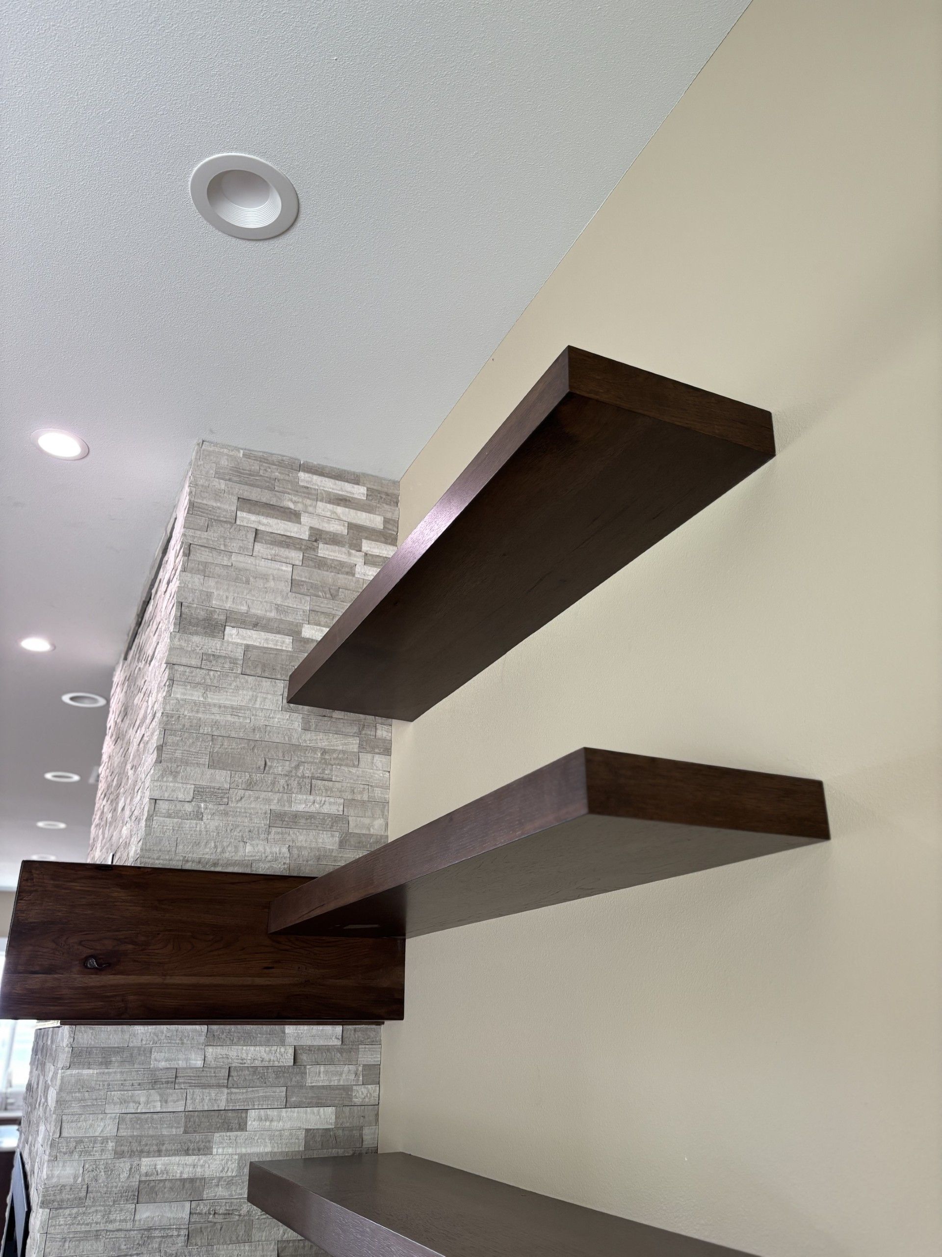 Three dark wooden floating shelves against a cream-colored wall with a stone accent. Recessed lighting is visible.