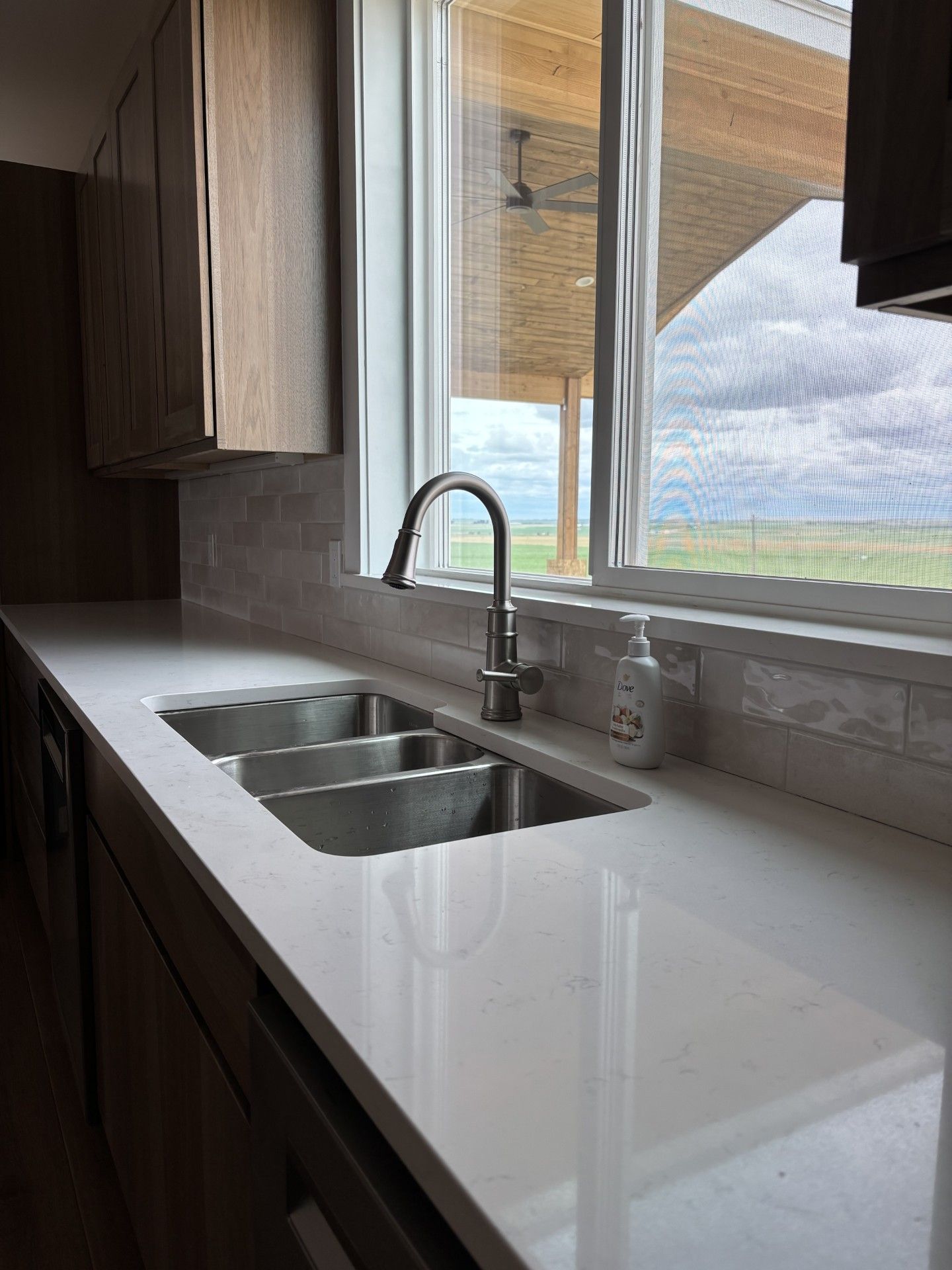 Kitchen with a white countertop, a triple-basin sink, and a window overlooking a landscape. Brown cabinets flank the sink.